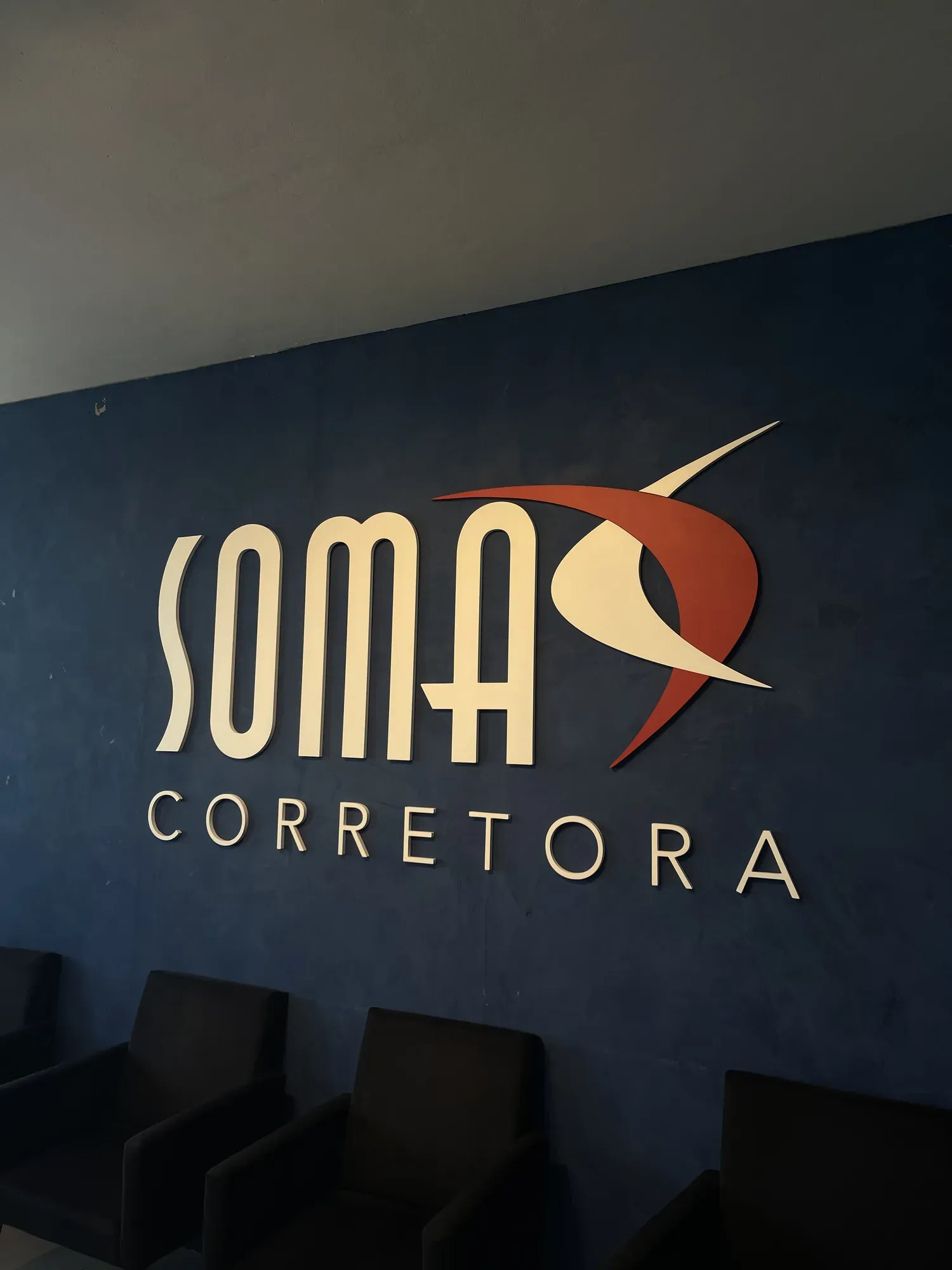 Recepção da Somax Corretora: o logo em relevo sobre a parede azul, com poltronas de atendimento à frente.