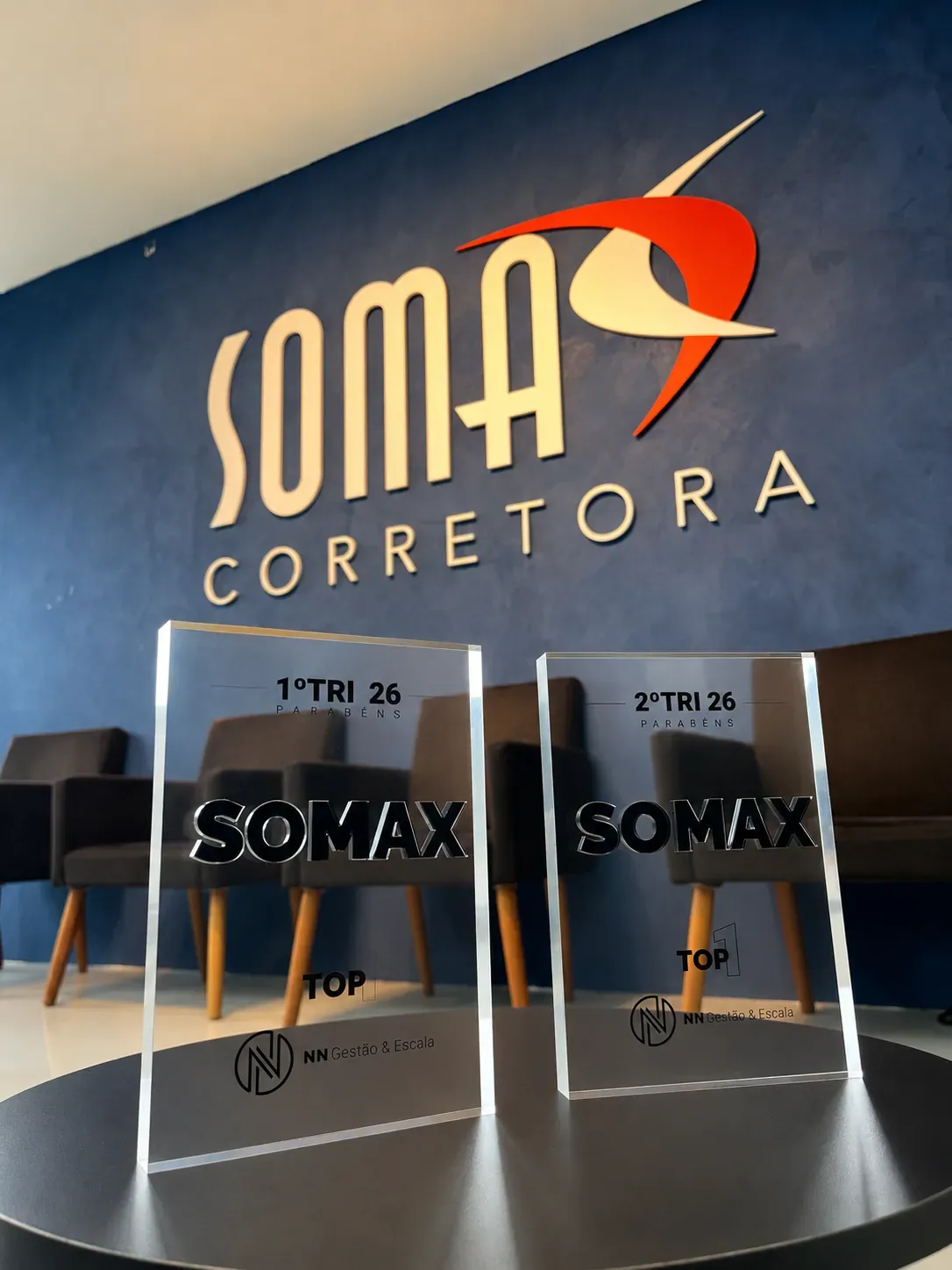 Dois troféus Somax de Top 1 em performance, referentes ao 1º e ao 2º trimestre de 2026, concedidos pela NN Gestão & Escala, sobre a mesa da recepção da corretora.